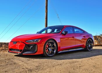 A red 2025 Audi RS e-tron GT Performance electric sports car is parked on a dirt road under a clear blue sky, with utility poles and wires in the background.