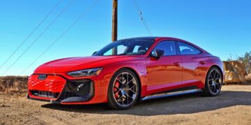 A red 2025 Audi RS e-tron GT Performance electric sports car is parked on a dirt road under a clear blue sky, with utility poles and wires in the background.
