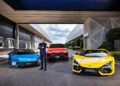 A man in a suit stands with arms crossed between a blue car, a red SUV, and a yellow sports car—reminiscent of Lamborghini’s strong Q3 results and record margins in the hybrid era—parked outside a modern building.