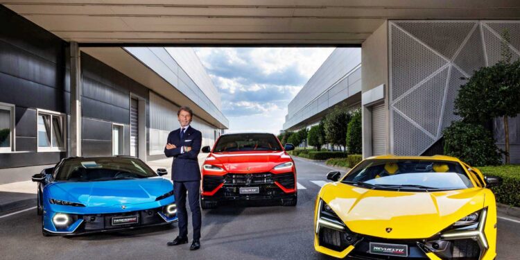 A man in a suit stands with arms crossed between a blue car, a red SUV, and a yellow sports car—reminiscent of Lamborghini’s strong Q3 results and record margins in the hybrid era—parked outside a modern building.