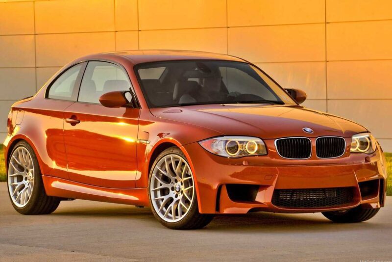 An orange E46 M3 coupe is parked outdoors in front of a beige wall, illuminated by sunlight, blending classic performance with modern-day style.