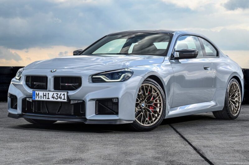 A silver BMW E46 M3 coupe with a sporty design and black accents is parked on a concrete surface under a cloudy sky, embodying modern-day performance in a classic disguise.