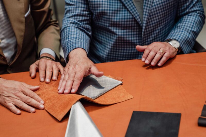 Two men in suits examine and touch fabric and material samples on a table, selecting options for the exclusive BOTTEGAFUORISERIE collection at the Alfa Romeo Maserati Launch.