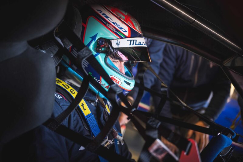 A race car driver wearing a helmet and racing suit sits inside an Alfa Romeo Maserati, secured with safety harnesses and a window net, preparing for the BOTTEGAFUORISERIE launch race.