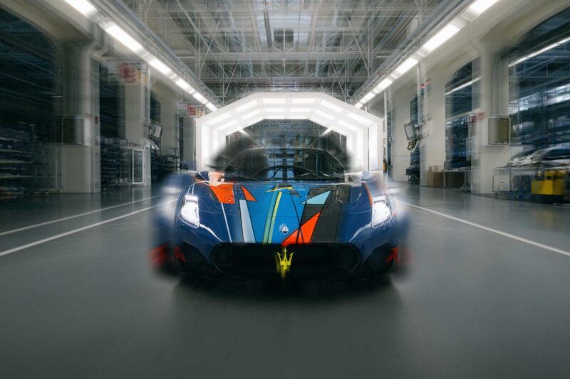 An Alfa Romeo with a colorful, geometric paint design is parked inside a brightly lit, modern automotive factory for the BOTTEGAFUORISERIE launch.