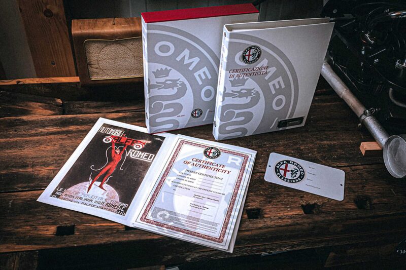 Two Alfa Romeo certification folders, a certificate of authenticity, a vintage advertisement, and an ID card displayed on a wooden table—perfect for any Alfa Romeo or Maserati enthusiast and ideal for BOTTEGAFUORISERIE collectors.