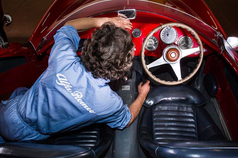 Person in a blue Alfa Romeo jacket sits in a vintage red car, reaching for the dashboard and steering wheel, seen from above—capturing the excitement of an Alfa Romeo Maserati Launch.