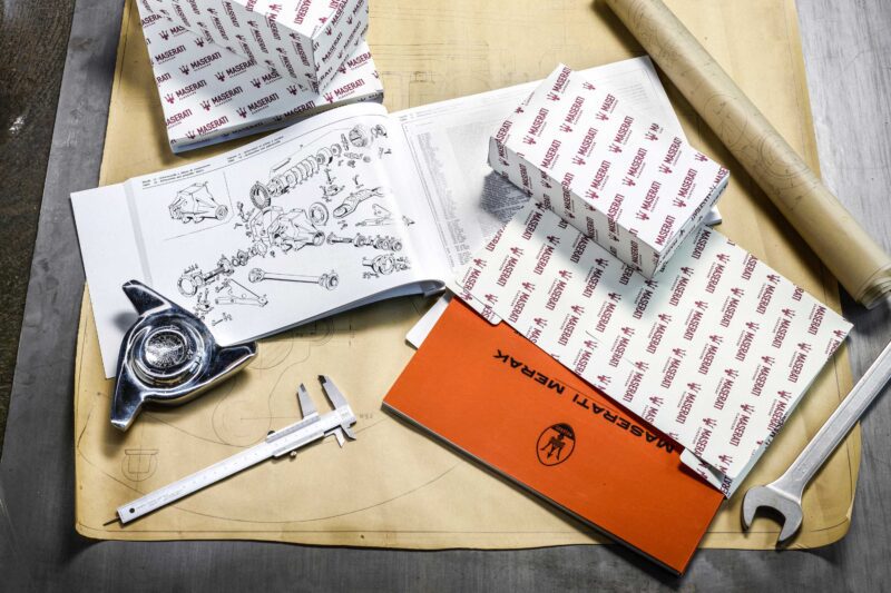 A table displays Alfa Romeo and Maserati car parts boxes, an exploded view manual, a vernier caliper, a wrench, a blueprint, and a classic car hubcap—capturing the spirit of BOTTEGAFUORISERIE craftsmanship.