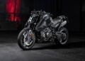 A matte black BRABUS 1400 R Signature Edition sport motorcycle is parked indoors on a concrete floor, with a dim background and red light illuminating the wall behind it, hinting at its exclusive 2026 allure.