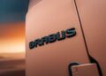 Close-up of a "BRABUS" badge on a matte orange vehicle, highlighting the bold style as Brabus debuts 800 Cabrio and XL 800 Cabrio, with a blurred sunset or dusk sky in the background.