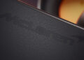 Close-up of a black leather surface embossed with the McLaren logo, with a blurred background featuring orange and metallic tones, reminiscent of the premium style found on Bowers & Wilkins Px8 S2 McLaren Edition Headphones.