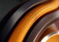 Close-up of an orange braided cable or hose, reminiscent of the Bowers & Wilkins Px8 S2 McLaren Edition headphones, encased in a metallic structure, with a textured black surface partially visible in the background.