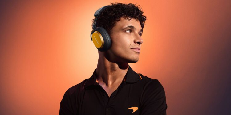 A young man wearing large over-ear Bowers & Wilkins Px8 S2 headphones and a black polo shirt with an orange logo stands against an orange gradient background.
