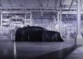 A covered car sits in the center of a modern, brightly lit automotive factory with advanced machinery in the background, hinting at the future of Bentley hybrids and electric vehicles.