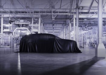 A covered car sits in the center of a modern, brightly lit automotive factory with advanced machinery in the background, hinting at the future of Bentley hybrids and electric vehicles.
