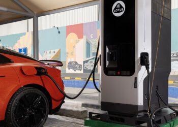 An orange electric car is charging at a Lotus-branded charging station under a canopy, with a colorful mural in the background.