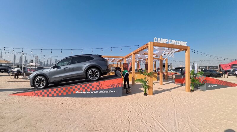 Feature Exclusive: 2025 Icons of Porsche Dubai—a silver SUV is displayed on an angled ramp under a wooden "Camp Cayenne" structure at an outdoor event on sandy ground.