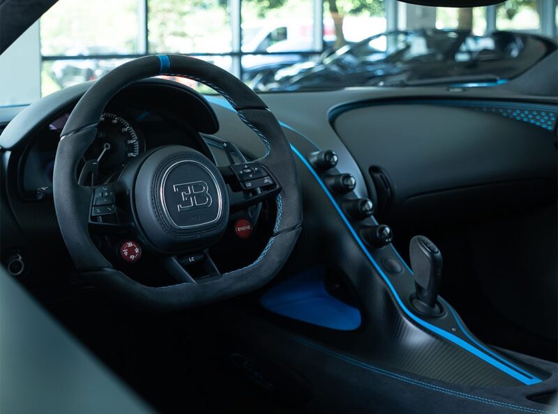 Close-up view of a Bugatti Chiron Pur Sport interior showing the steering wheel, dashboard, gear selector, and blue accents; experience luxury and precision—this stunning model is now for sale.