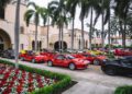 A row of red and yellow sports cars parked outside a large, elegant building with palm trees and flower beds nearby.