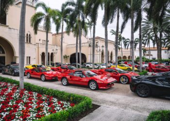 A row of red and yellow sports cars parked outside a large, elegant building with palm trees and flower beds nearby.