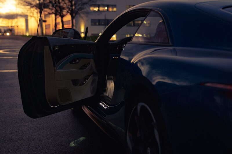 A dark blue Bentley sports car with its driver door open, parked outside at dusk near a building with illuminated windows and bespoke welcome lamps.