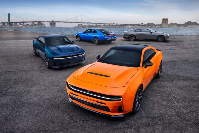 Four modern muscle cars in orange, green, blue, and gray—each as striking as a powerful sedan—are parked on a paved lot near a river, with a bridge and city skyline in the background.