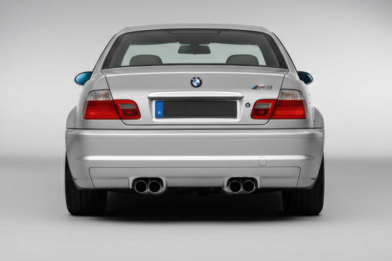 Rear view of a silver E46 M3 sports car, showing dual exhaust pipes, taillights, and a European license plate against a plain background—a modern-day classic in no disguise.