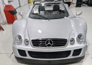 A silver Mercedes-Benz CLK-GTR Roadster convertible sports car, valued as a $13M car, is parked indoors on a clean, white floor with its top down and front view visible.