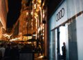 A man in a suit exits a glass-fronted building displaying the Audi logo at night, during what appears to be the Audi Concept C’s London debut, as people walk along a brightly lit city street.