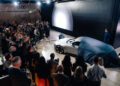 A crowd gathers around as a silver Audi Concept C, partially covered by a blue cloth, makes its London debut at an indoor event with a presenter standing nearby.