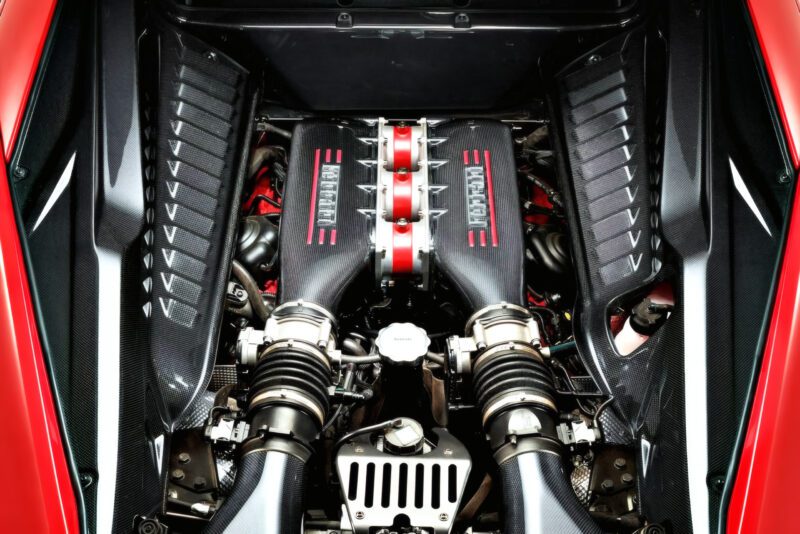 Close-up view of a car engine with visible air intake components and "Ferrari" branding, showcasing the last naturally aspirated mid-engine V8 Ferrari ever built, surrounded by striking black and red engine bay detailing.