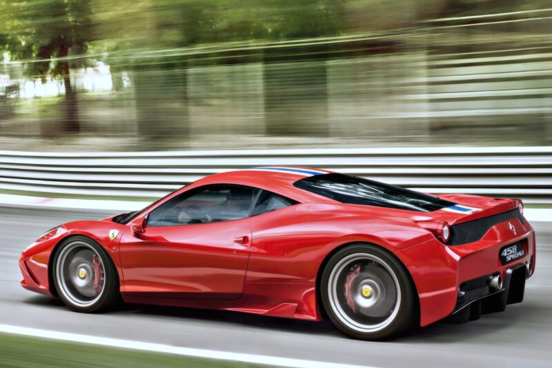 A red Ferrari 458 Speciale—the last naturally aspirated mid-engine V8 Ferrari ever built—drives at speed on a racetrack, blurred trees and barriers streaking past in the background.