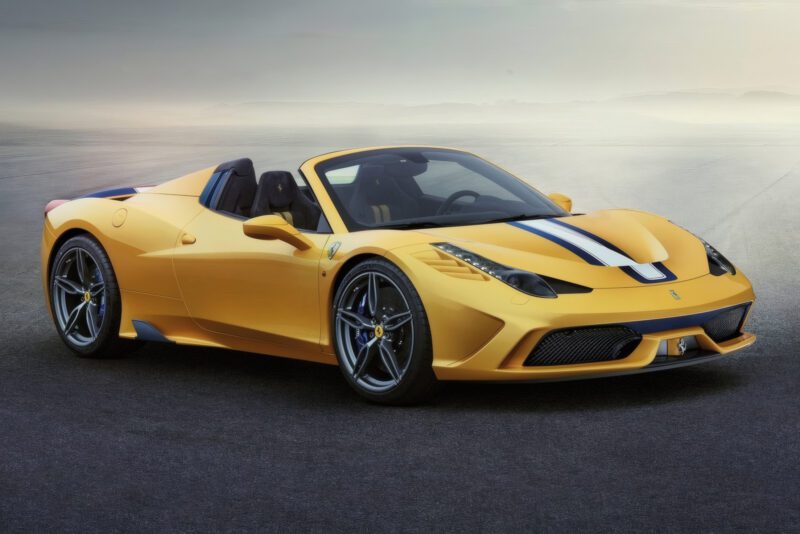 A yellow convertible sports car, known as the last naturally aspirated mid-engine V8 Ferrari ever built, with blue and white racing stripes is parked on an empty paved surface under an overcast sky.