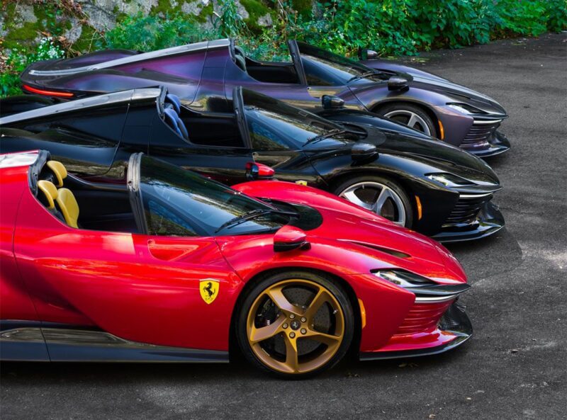 Three ultra-rare Ferrari Daytona SP3 convertible sports cars in red, black, and dark purple are parked side by side on a paved surface near greenery, showcased by Miller Motorcars.