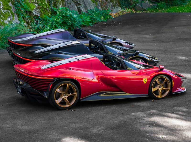 Three luxury sports cars, including an ultra-rare Ferrari Daytona SP3 with gold wheels in the foreground, are parked in a row on a paved surface near greenery and a rock wall at Miller Motorcars.