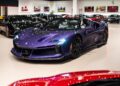 A Violetto Dino Ferrari SF90 Spider is displayed at Ferrari Lake Forest's luxury showroom, surrounded by other high-end vehicles under bright lighting.