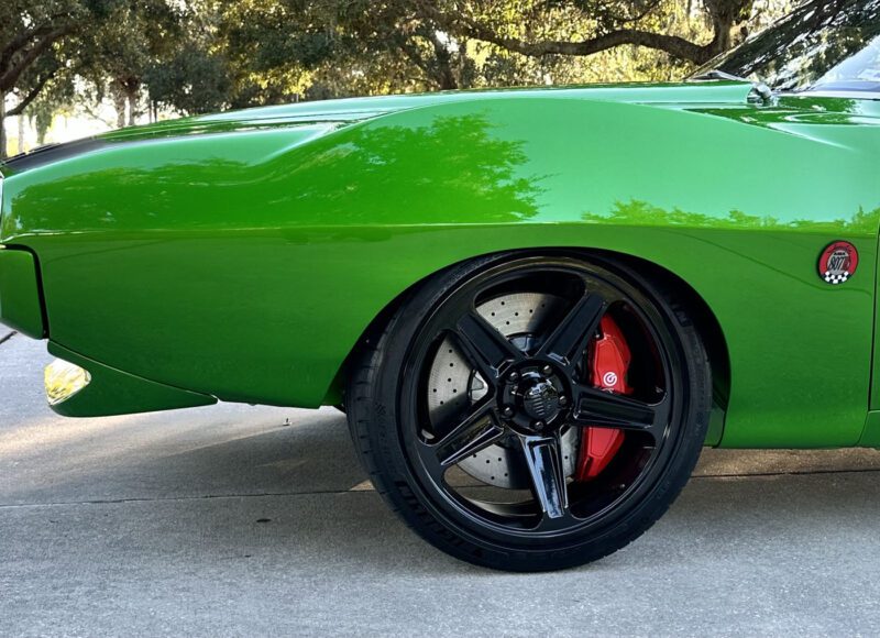 Close-up of a bright green Pro Touring AMC Javelin’s front left wheel, featuring a black rim, red brake caliper, and Hellcat logo on the fender, with trees and pavement in the background.
