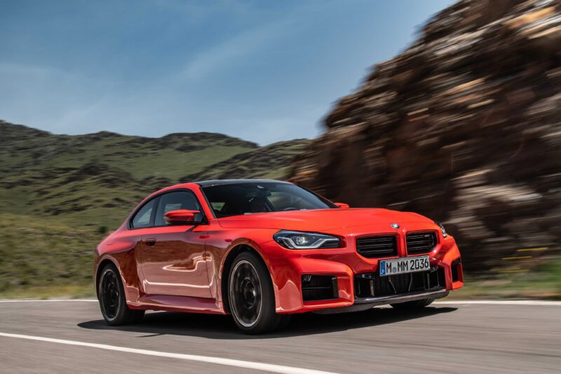 A red E46 M3 BMW coupe drives on a paved road, its modern-day design in perfect disguise against the blurred cliffs and greenery in the background.