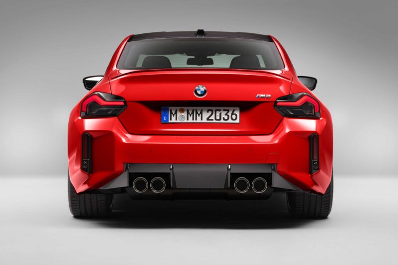 Rear view of a red BMW M2 sports car, its quad exhaust pipes and badges hinting at modern-day performance, yet echoing the spirit of the E46 M3—no disguise here, just pure driving passion against a neutral background.