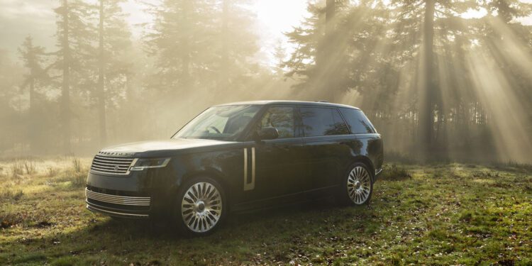 A black Range Rover SUV parked on grass in a sunlit forest clearing with rays of sunlight streaming through the trees.