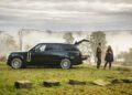 A black SUV with its trunk open is parked on grass near rocks. Two people stand nearby, talking, with foggy trees and a cloudy sky in the background.