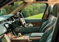 The image shows the luxurious front interior of a Range Rover with green and tan leather seats, wood trim, and advanced controls, parked near a grassy, tree-lined landscape—a signature touch from Holland & Holland.