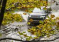 A dark-colored Range Rover SUV drives through a shallow, muddy water crossing, with splashes around its wheels and autumn leaves in the foreground.