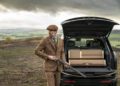 A man in a brown checkered suit holds a shotgun next to an open Overfinch Range Rover trunk containing gun cases, standing outdoors on a cloudy day.