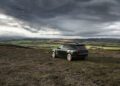 A dark green Range Rover is parked on a grassy hill overlooking a wide countryside landscape under a cloudy sky.