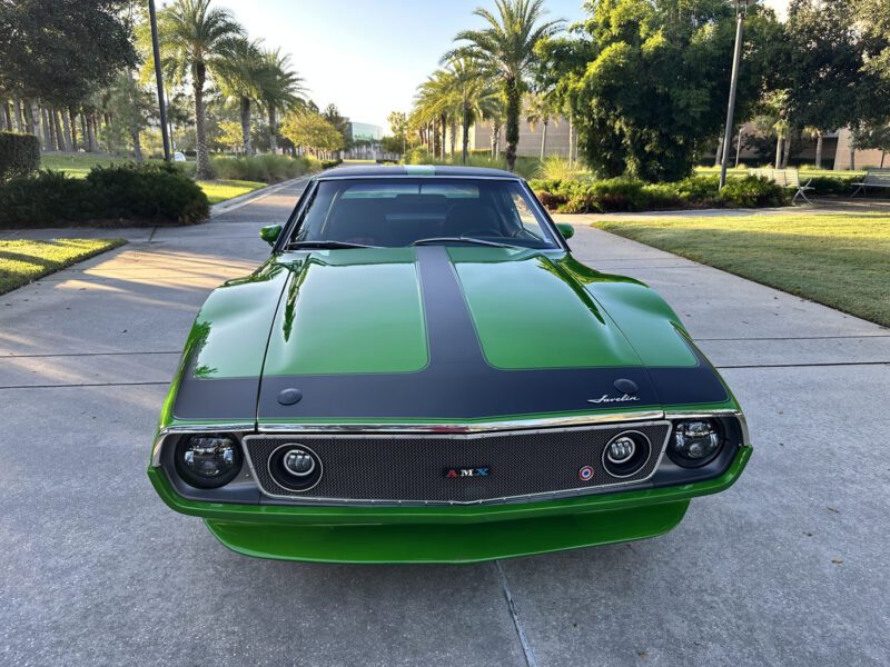 A 1972 AMC Javelin AMX, dubbed "Green Lightning" for its striking color and Pro Touring style, is parked on a sunlit driveway lined with palm trees, flaunting sleek black hood detailing.