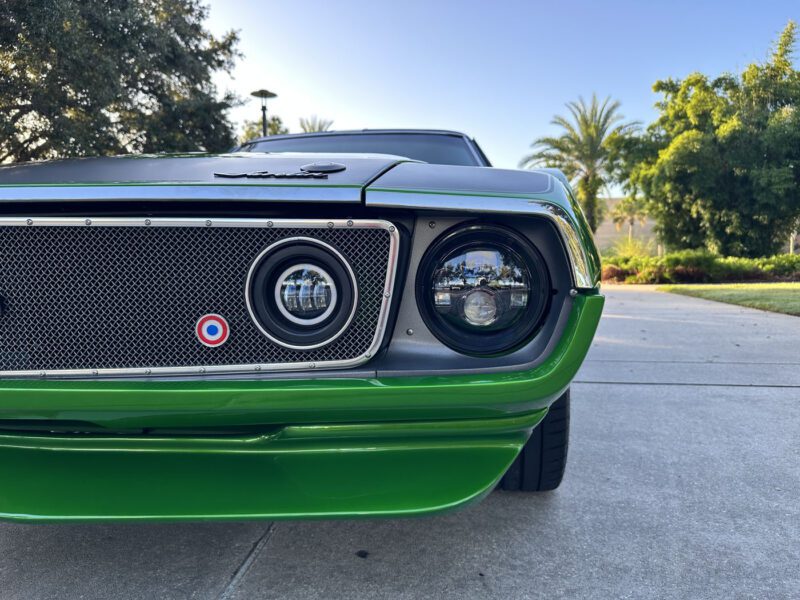 Close-up view of the front left side of a green 1972 AMC Javelin Pro Touring parked on a driveway, showing the grille, headlight, and sleek bodywork. Trees and clear sky are visible in the background.