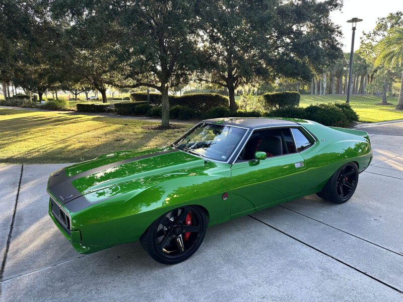 A green 1972 AMC Javelin muscle car with black accents, known as "Green Lightning," is parked on a driveway, surrounded by grass, trees, and landscaping on a sunny day.