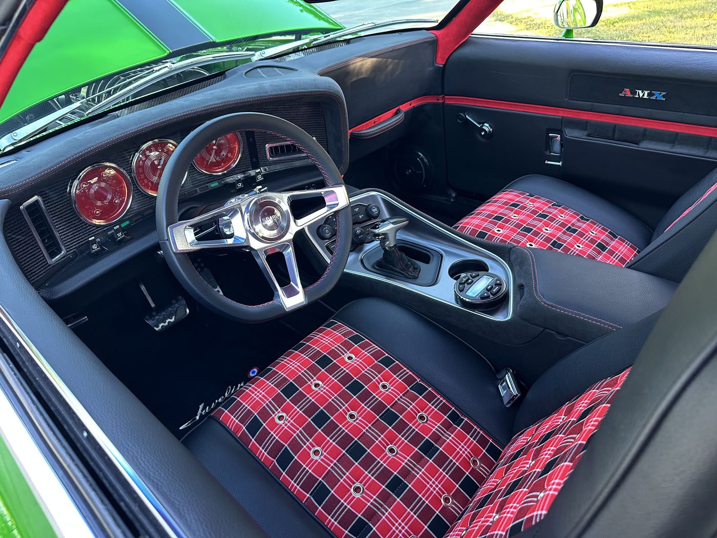 Interior of a 1972 AMC Javelin Pro Touring featuring a black dashboard with red gauges, custom plaid red and black seat upholstery, and a modern steering wheel and center console.