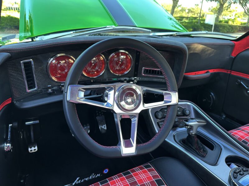 Close-up view of a 1972 AMC Javelin's interior showing a three-spoke steering wheel, red-accented dashboard with round gauges, and plaid seat upholstery, capturing classic muscle car style.